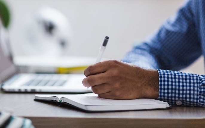 Close up photo of a man's hand writing in a notebook with a laptop in the background.